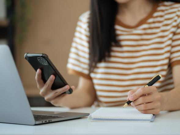 A young woman sits in front of a laptop, holding a smartphone and making notes on a pad