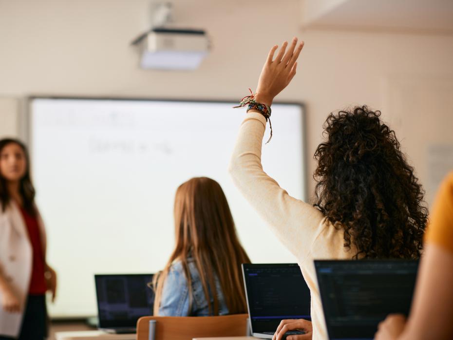 A class full of students from behind, with one raising their hand to answer a question