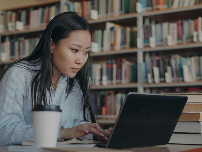 Female Asian college student writing in library on a laptop