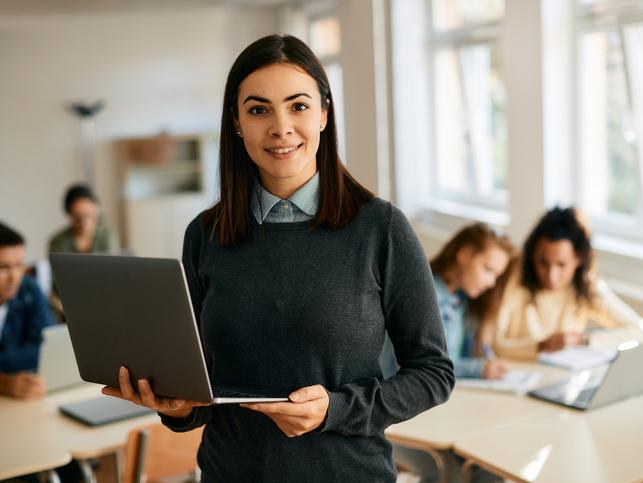 A woman stands in front of a class, holding a laptop