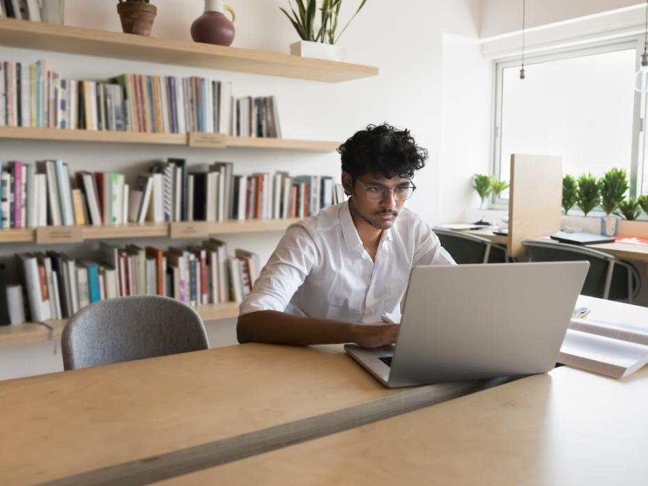 A male researcher writing at his laptop surrounded by shelves of books