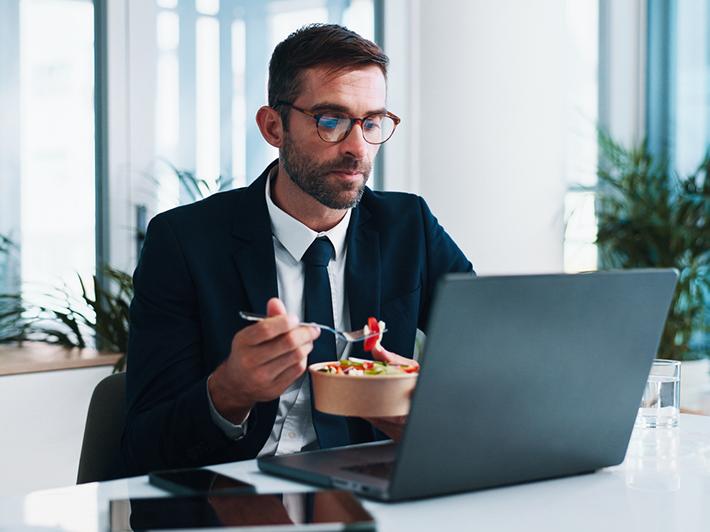 Businessman doing online study at laptop while eating takeaway lunch