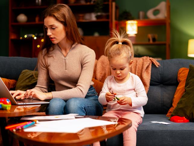 A mother works at a laptop while entertaining a small child