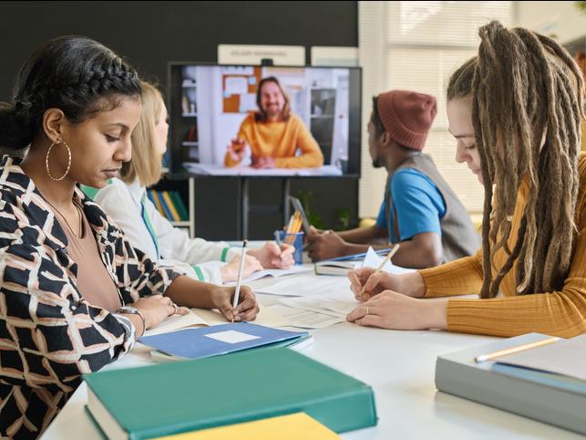 Students in a classroom with an online student appearing on a screen at the end of the table