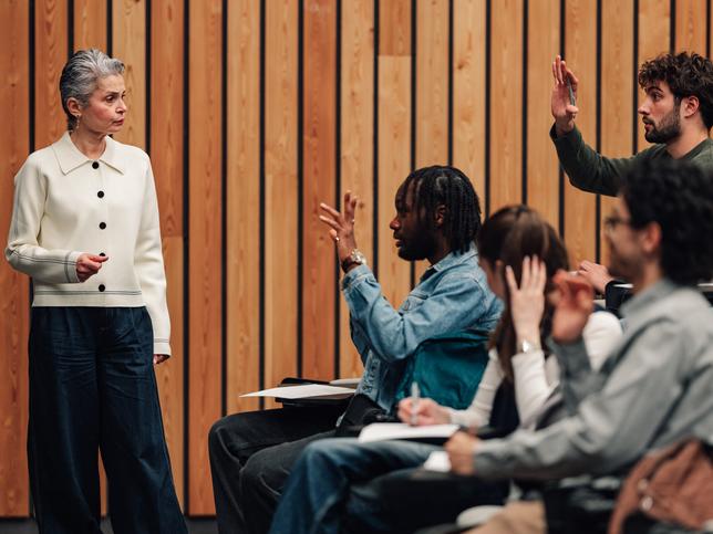 A professor explaining course structure to a class full of students, some of whom have their hands raised