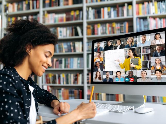 A female student taking notes from her online class with other students and a teacher appearing in a gallery view on her screen