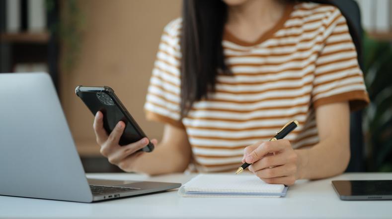 A young woman sits in front of a laptop, holding a smartphone and making notes on a pad