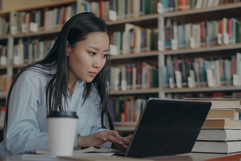 Female Asian college student writing in library on a laptop