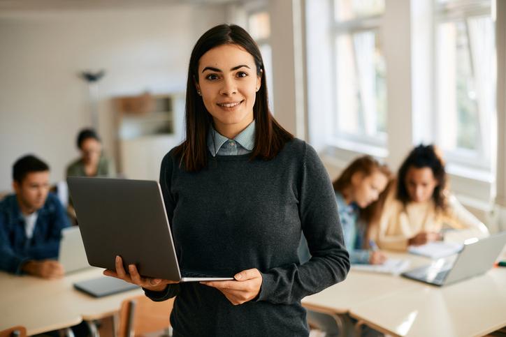 A woman stands in front of a class, holding a laptop