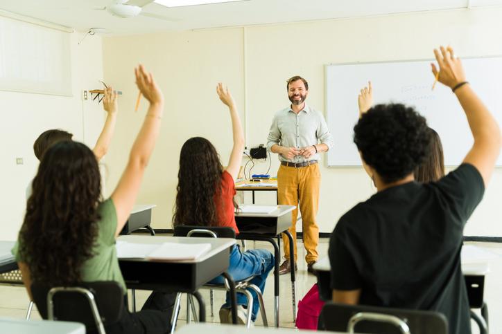 A teacher smiles as students raise their hands en masse