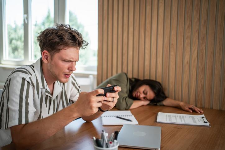 A young academic plays a game on his phone while his co-worker naps on the desk