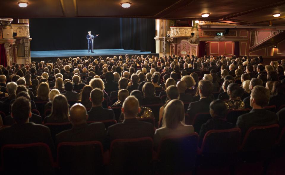 An audience watching a man on stage in a theatre