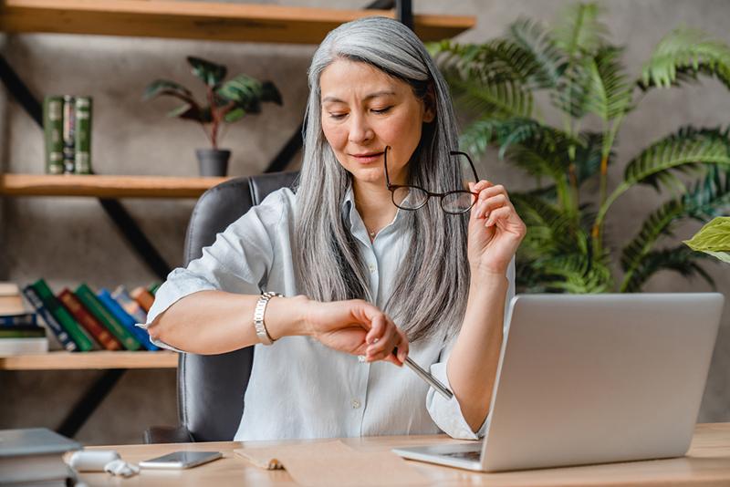 Mature Asian woman looking at watch with laptop