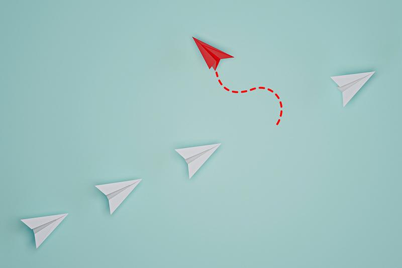 red paper airplane leaving line of white paper airplanes