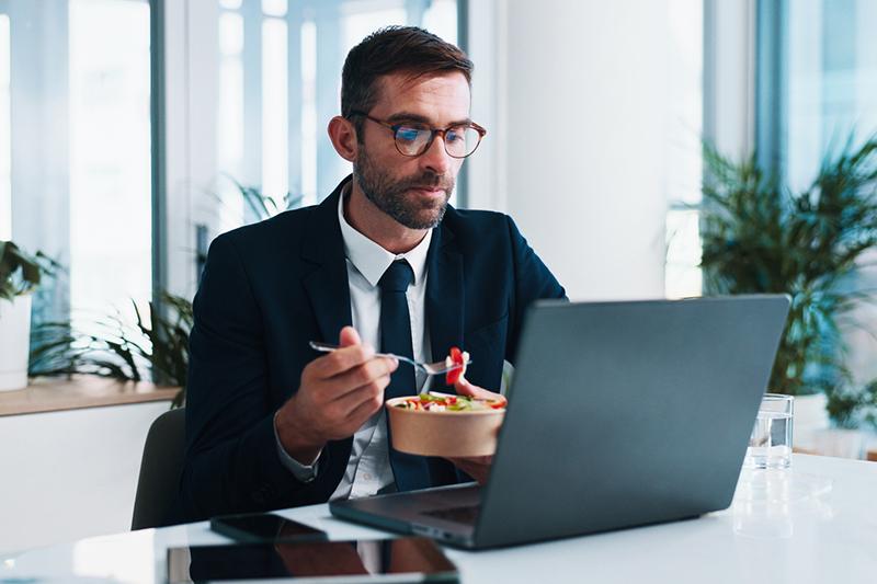 Businessman doing online study at laptop while eating takeaway lunch