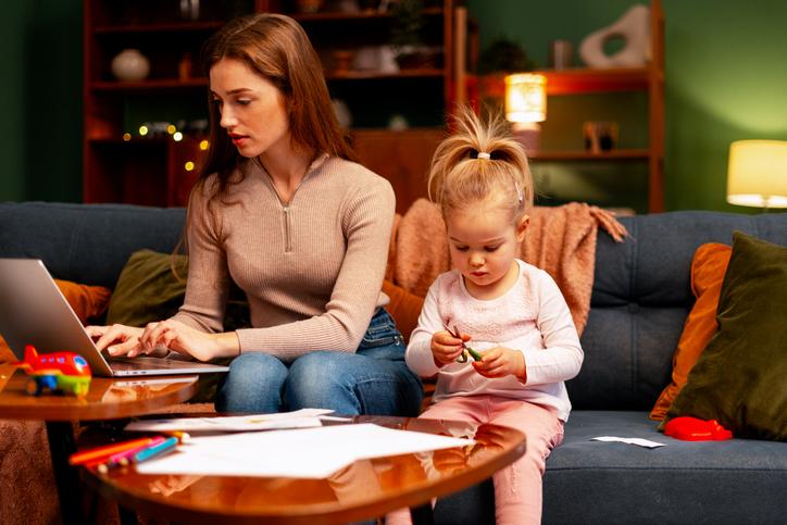 A mother works at a laptop while entertaining a small child