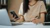 A young woman sits in front of a laptop, holding a smartphone and making notes on a pad