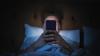 A man sits in darkness, illuminated by a smartphone that blocks his face from view