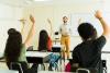 A teacher smiles as students raise their hands en masse